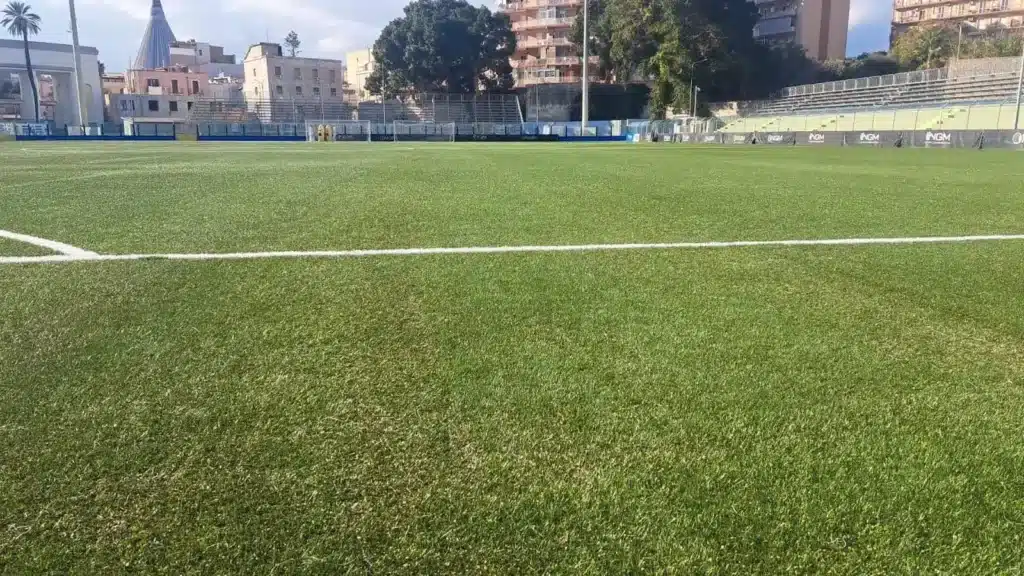 Veduta in primo piano del manto erboso sintetico dello stadio Nicola De Simone di Siracusa. Sullo sfondo si notano le tribune, i palazzi della città e la guglia del Santuario della Madonna delle Lacrime sotto un cielo parzialmente nuvoloso.